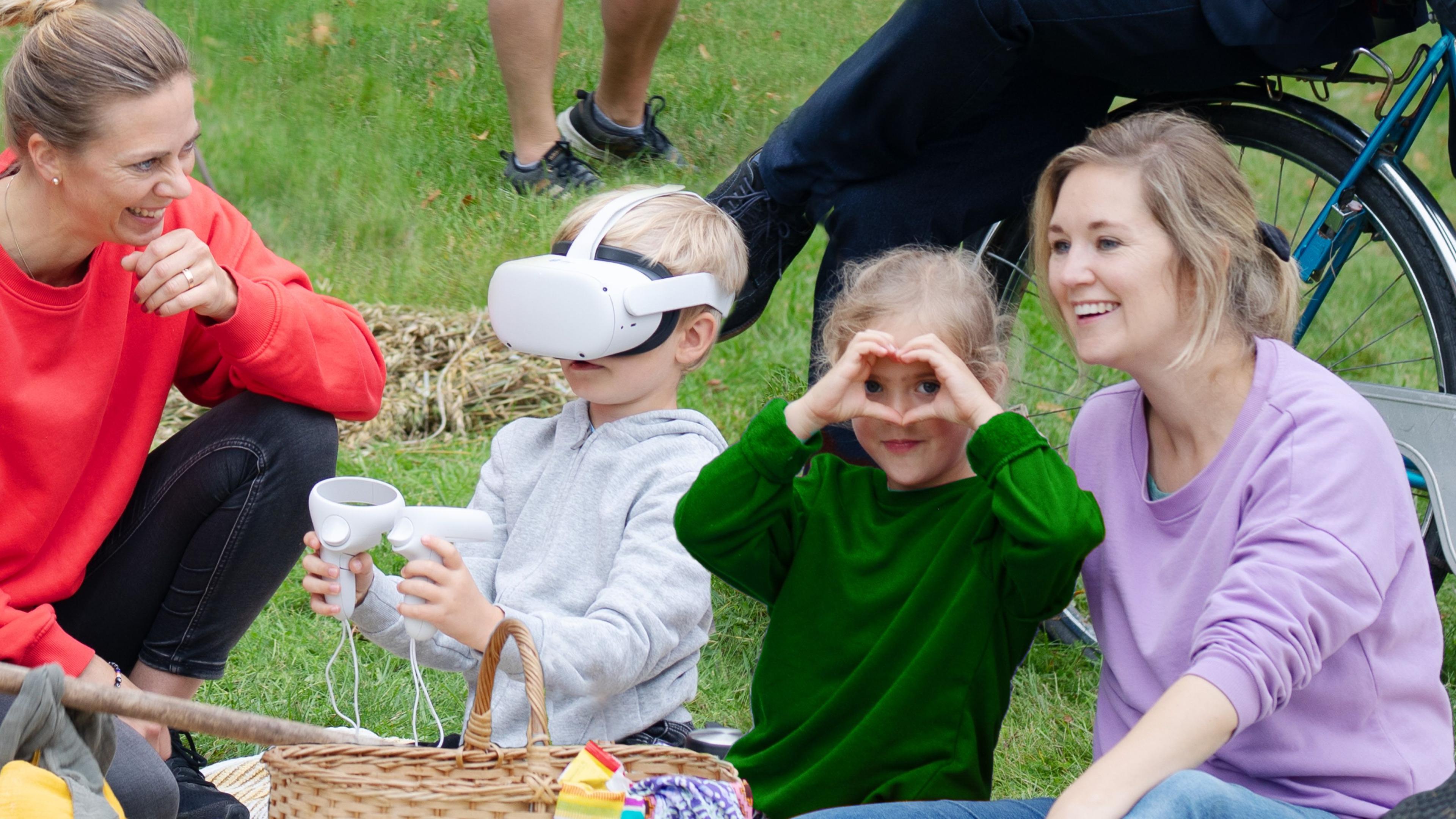 Två föräldrar och två barn sitter på gräsmatta där ett barn har på sig VR glasögon
